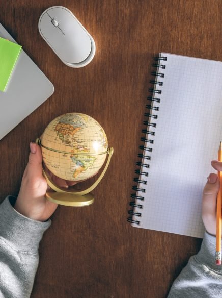 A globe in the hands of a teacher, a laptop and a notepad on the background of a wooden table. School and education concept, top view.