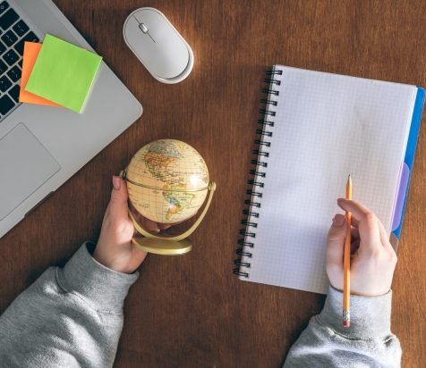 A globe in the hands of a teacher, a laptop and a notepad on the background of a wooden table. School and education concept, top view.